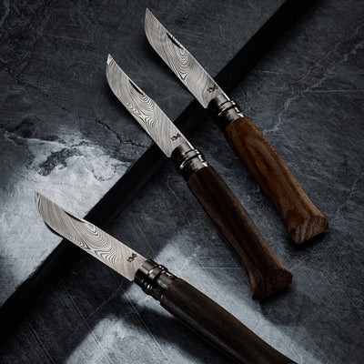 Three folding knives with patterned Damascus steel blades and dark brown wooden handles arranged diagonally on a dark textured slate surface.