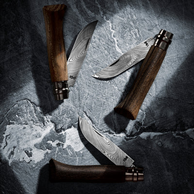 Three folding pocket knives with dark wood handles and patterned Damascus steel blades, arranged on a dark textured stone surface with spotlight in the center.