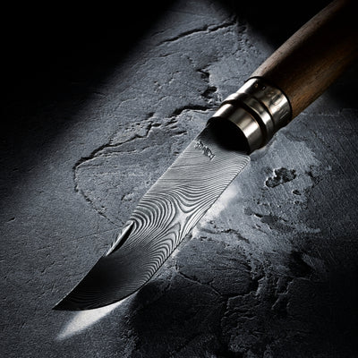 A close-up of a Damascus steel knife with a wavy pattern blade, a silver bolster, and a wooden handle, resting on a dark textured stone surface under dramatic spotlighting.