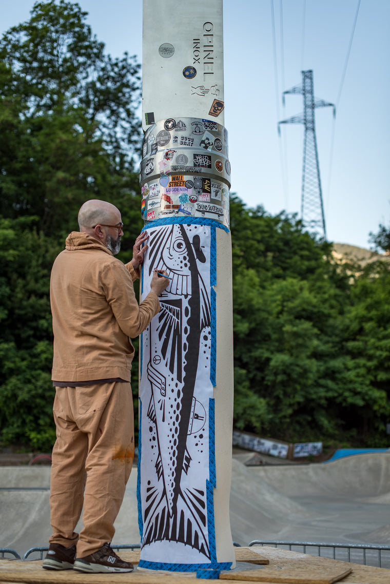 A man in a tan hoodie and sweatpants stands beside a tall concrete pole outdoors, applying a large black-and-white fish mural while blue painter's tape wraps the lower section. The pole is covered in stickers near the top, with trees, a hill, and a power line tower in the background.