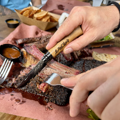A close-up lifestyle shot of a person carving a cooked ribeye steak with a knife featuring a light wood handle engraved with 'LeRoy & Lewis'.
