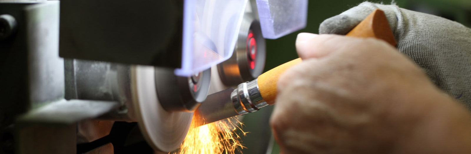 A close-up action shot of a person wearing a gray work glove and holding a wood-handled tool while operating a metal grinder, with bright orange sparks flying.