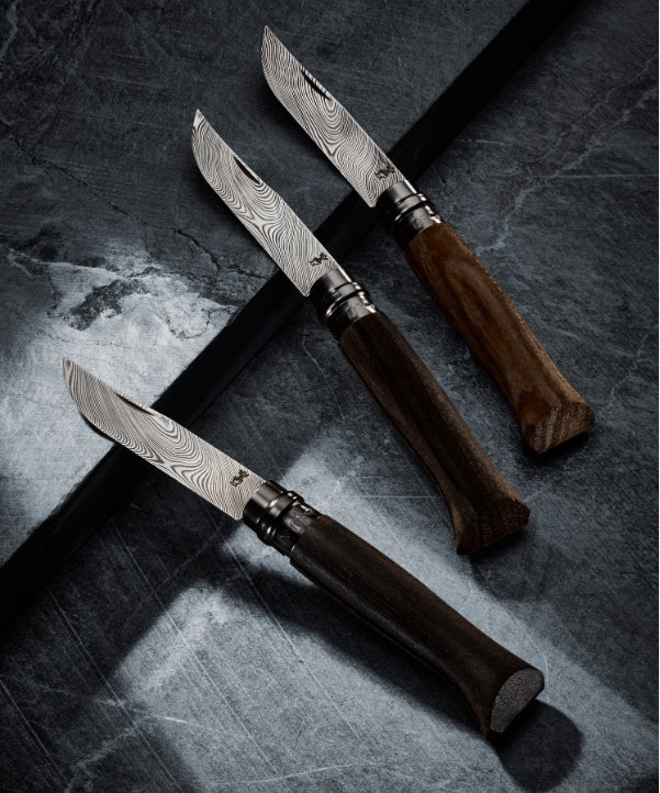 Three folding knives with patterned Damascus steel blades and dark wooden handles arranged diagonally on a dark textured slate surface.