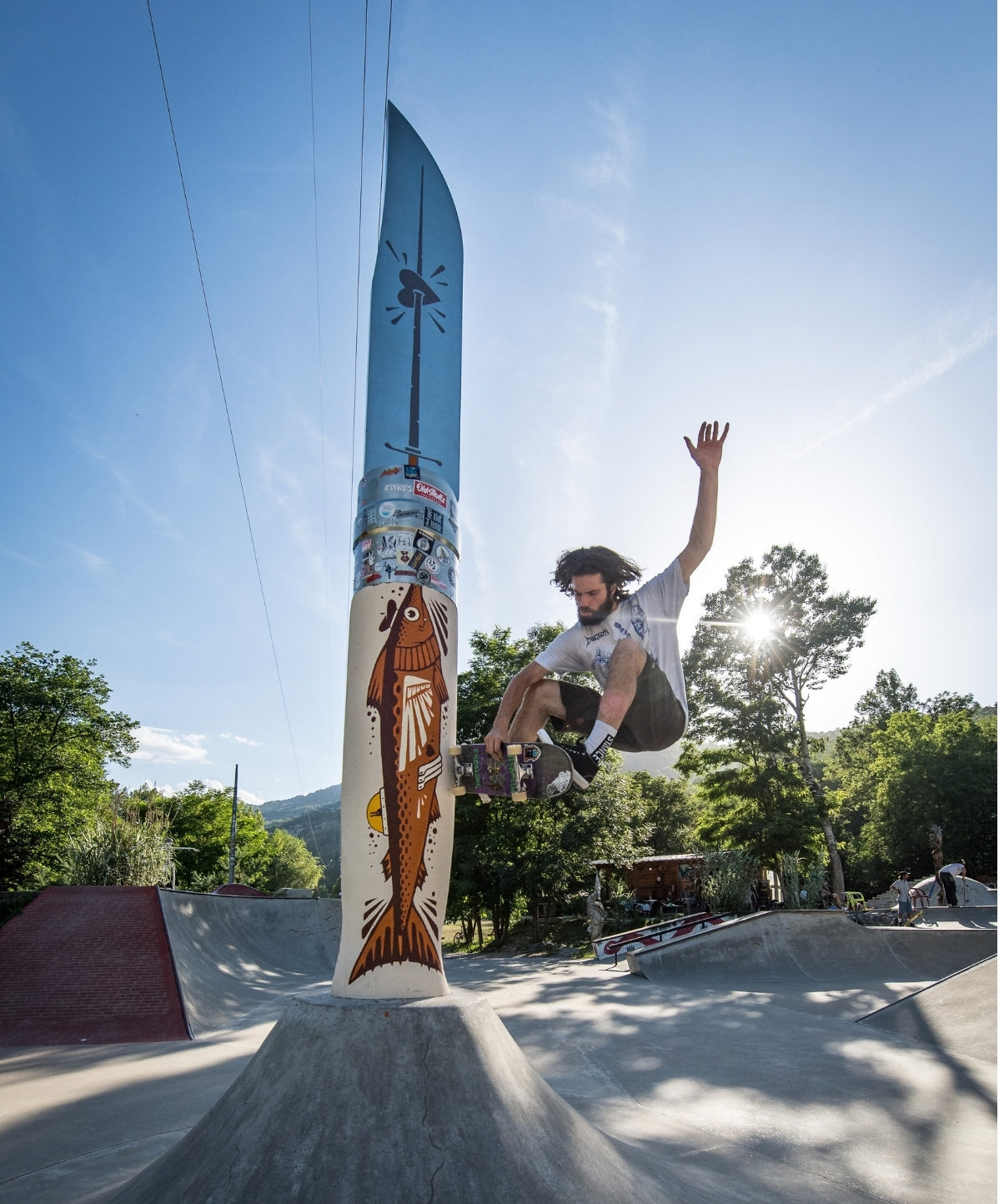 A skateboarder in mid-air performs a trick beside a tall painted giant knife at an outdoor skatepark under a bright blue sky with trees in the background.