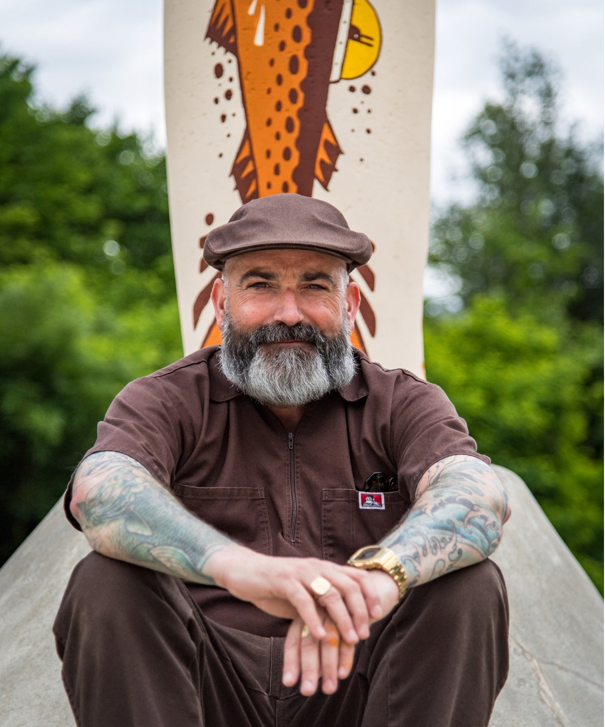 A man with a gray beard and tattoos sits outdoors in front of a tall painted column. He is wearing a brown flat cap, a brown short-sleeve button-front shirt, and brown pants.