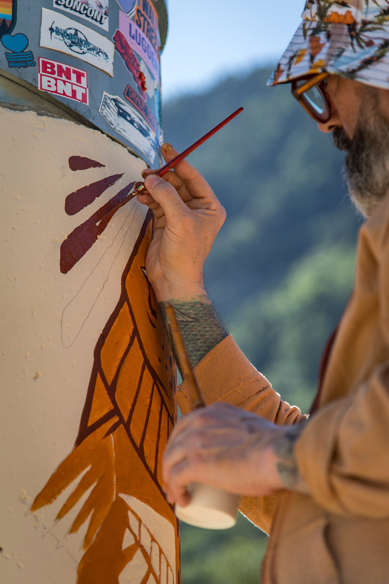 A bearded man wearing a patterned cap and glasses paints a mural on a pillar outdoors. The pillar is decorated with stickers at the top and features a painted design in orange, brown, and white.