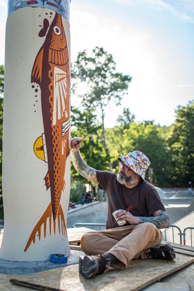A bearded man with tattoos sits on a wooden board outdoors, painting a large brown fish mural on a white cylindrical column. The background shows a sunny outdoor park or skate area with trees and a clear blue sky.