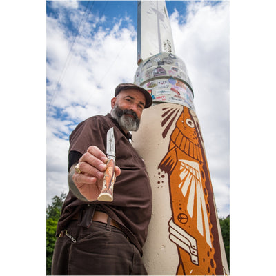 A bearded man wearing a brown shirt and cap holds up a folding knife with a natural wood handle toward the camera, standing next to a tall metal pole covered in stickers and featuring a large orange fish mural under a blue sky.
