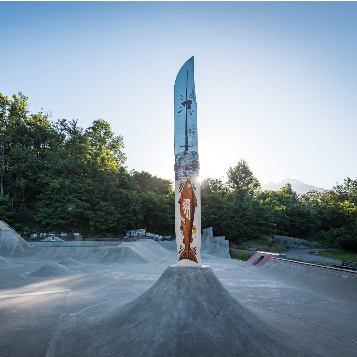 A tall decorative knife-shaped sign or sculpture stands centered on a concrete ramp at an outdoor skatepark, with trees, hills, and a blue sky in the background. The sign has a silver blade and a cream-colored handle decorated with a brown fish illustration and small red and yellow details.