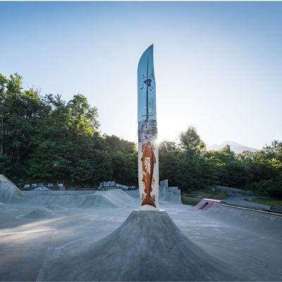A tall decorative knife-shaped sign or sculpture stands centered on a concrete ramp at an outdoor skatepark, with trees, hills, and a blue sky in the background. The sign has a silver blade and a cream-colored handle decorated with a brown fish illustration and small red and yellow details.