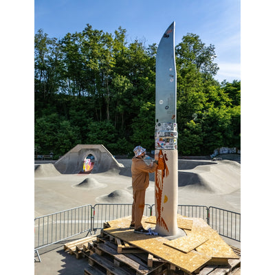 A person in a tan outfit stands on a wooden platform at an outdoor skatepark, adjusting a very tall, narrow metallic silver sculpture or pole. The sculpture has stickers and colorful painted details near the base.