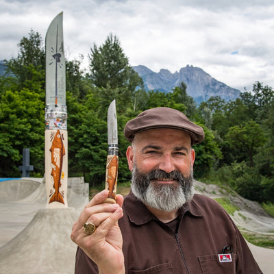 A smiling bearded man in a brown cap and brown shirt holds up a small decorative knife in front of a skatepark with large knife sculptures and mountains in the background.