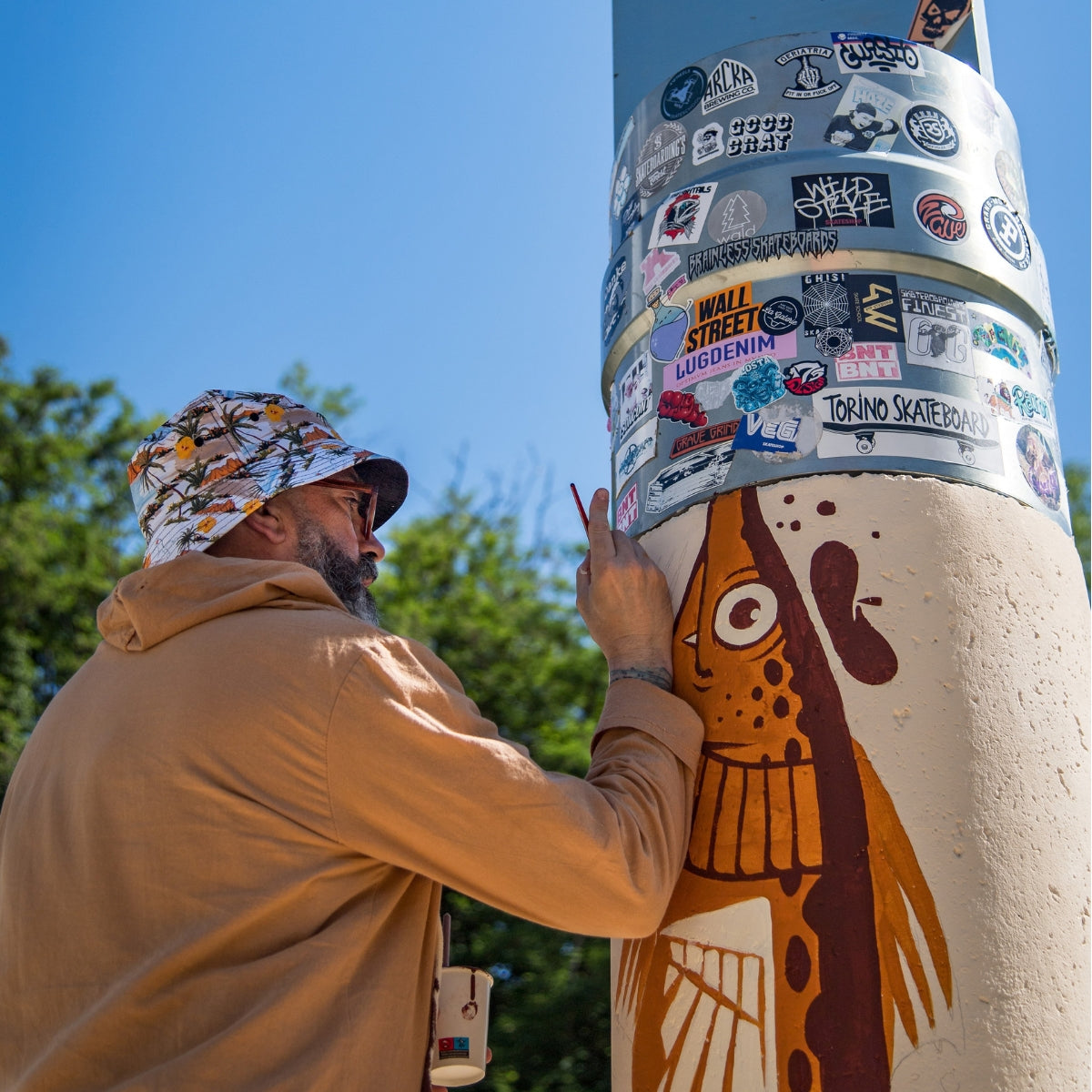 A man in a tan hoodie and camouflage bucket hat paints a mural on a concrete pole outdoors. The pole is covered in various stickers and features a painted fish design near the bottom.