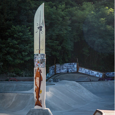 A large decorative knife displayed upright in a concrete skatepark, with a forest background and ramps visible behind it. The knife has a cream-colored upper section with a black sword illustration and a lower section painted with a brown fish and other small illustrations.