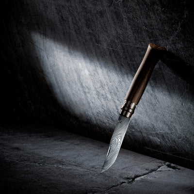 A folding knife in open position with a Damascus steel blade and a dark brown wooden handle, shown diagonally planted in the ground against a dark textured background with dramatic spotlighting.